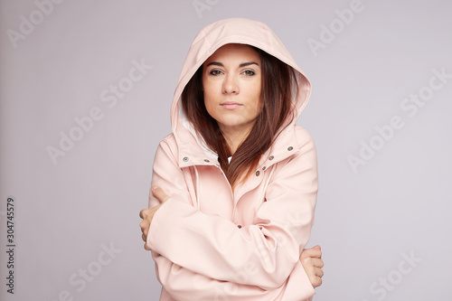 Gloomy young mixed race woman feels cold, looking at camera, frowning face, tries to warm herself, trembles after walking during heavy rain, keeps arms crossed over chest, wears pink raincoat.