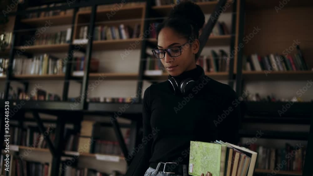 Cheerful african woman standing stepping down stairs in small library holding pile of books. Wearing black sweater, headphones on neck and stylish eyeglasses. Low angle footage Cheerful african woman standing stepping down stairs in small library holding pile of books. Wearing black sweater, headphones on neck and stylish eyeglasses. Low angle footage