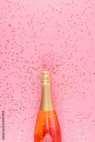 A bottle of pink champagne surrounded by gold confetti stars on a pink pastel background. © valkyrielynn