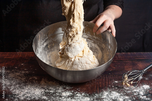 A cook in black kneads the dough in a metal bowl for cooking bread in the kitchen.