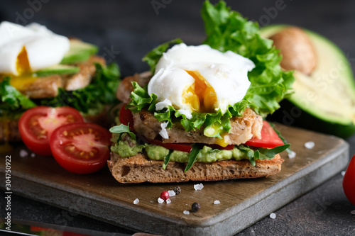 Delicious sandwich with avocado and poached egg, cherry tomatos and arugula on a dark background.