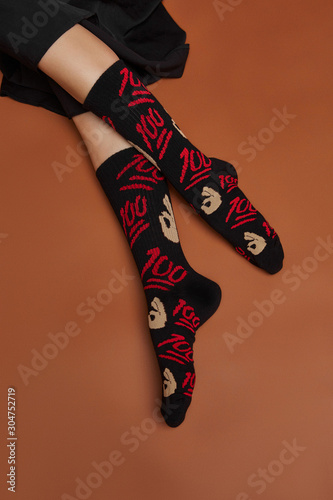 Cropped shot of a girl's foots, lying on a brown background. It is black artwork socks with lettering "100" and ok hand emoji print on foots. 