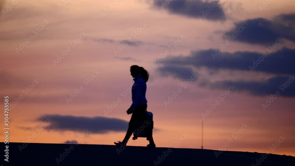 Woman walk with small suitcase at empty road, silhouetted shot against dusk sunset sky. Tired solo traveller after long way, girl limp slightly but still moving on Woman walk with small suitcase at empty road, silhouetted shot against dusk sunset sky. Tired solo traveller after long way, girl limp slightly but still moving on