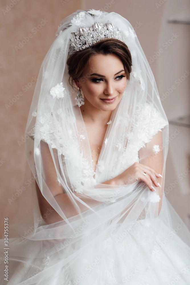 Wedding day. Bride portrait wedding makeup and hairstyle, fashion bride. Beautiful bride with veil. The girl hugs herself. Bride stands by the window and enjoys the sun's rays. Marriage. 