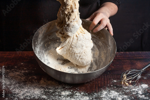 A cook in black kneads the dough in a metal bowl for cooking bread in the kitchen.