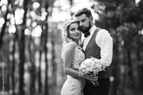 Sensual Black and white foto of bride and groom. Stylish couple of happy newlyweds posing in the park on their wedding day. Handsome bearded groom admires and kisses pretty bride. Together.