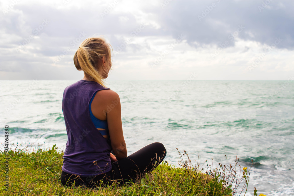 Young woman, blonde, is sitting on the grass and looks down on the sea. Relaxing after training 