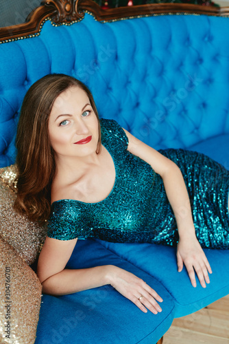 Beautiful woman in green dress sitting on blue velvet chesterfield sofa, copy space. Cozy home moment. Happy New Year. Christmas concept photo
