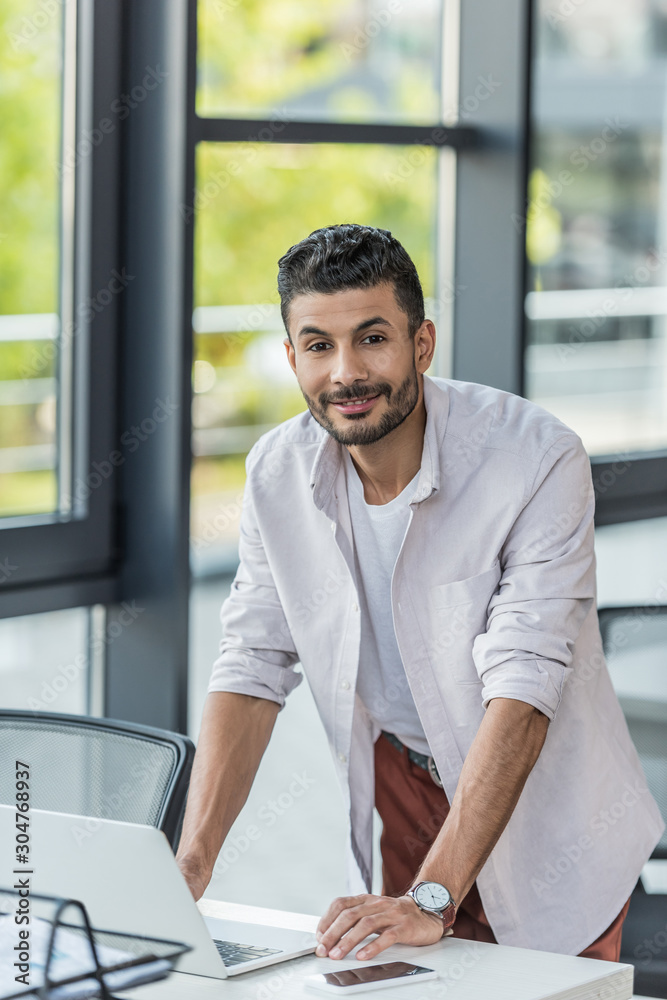 young businessman smiling at camera while standing near desk and using laptop young businessman smiling at camera while standing near desk and using laptop