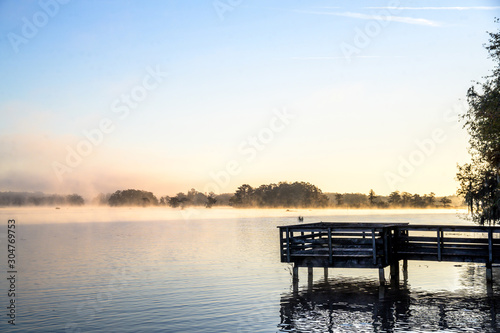 Misty Morning Lake with Autumn Colored Trees, Foliage, and Dock © Steven
