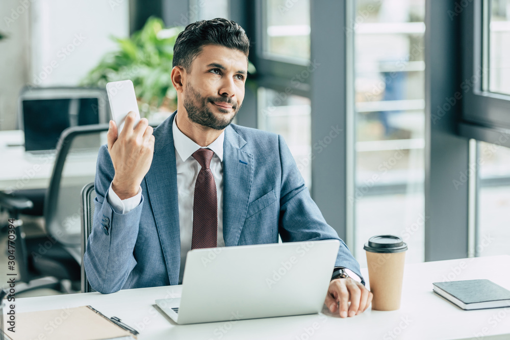 bored businessman looking away while sitting near laptop and holding smartphone bored businessman looking away while sitting near laptop and holding smartphone