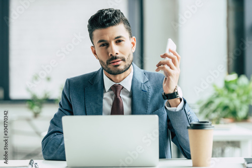 bored businessman looking at camera while sitting near laptop and holding smartphone