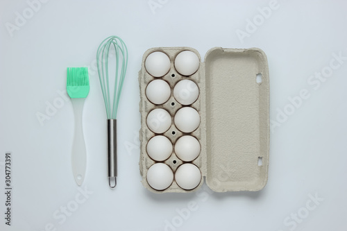 The concept of cooking. Cardboard tray of eggs  kitchen tools  whisk  brush  on white background. Top view