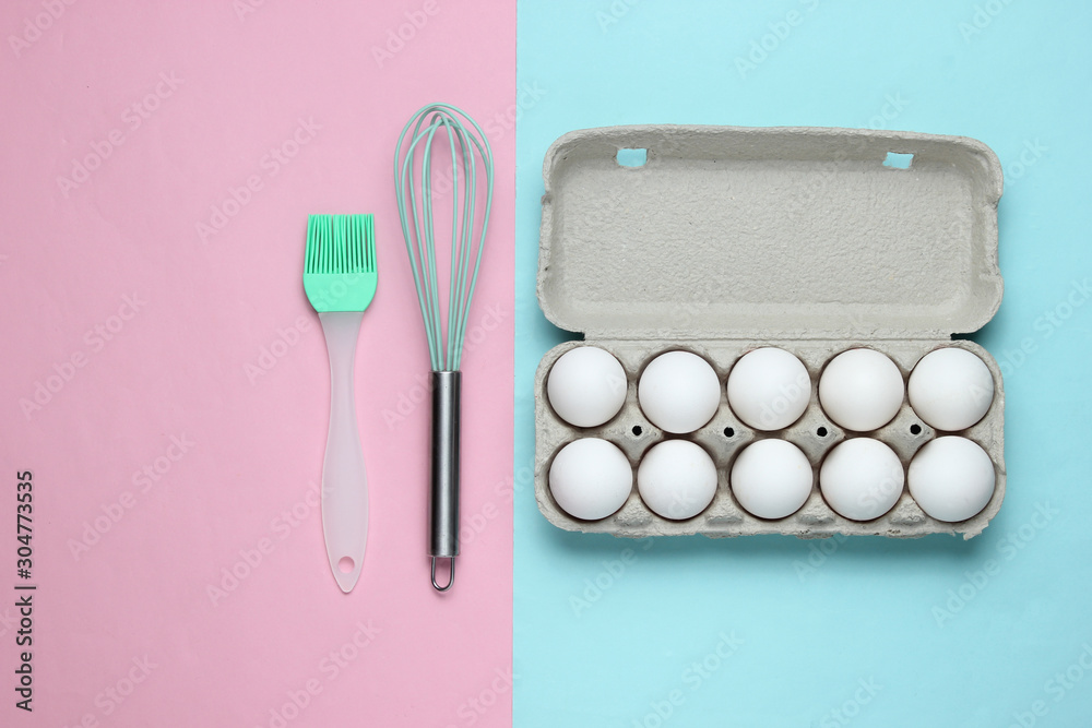 The concept of cooking. Cardboard tray of eggs, kitchen tools (whisk, brush) on a blue-pink pastel background. Top view The concept of cooking. Cardboard tray of eggs, kitchen tools (whisk, brush) on a blue-pink pastel background. Top view