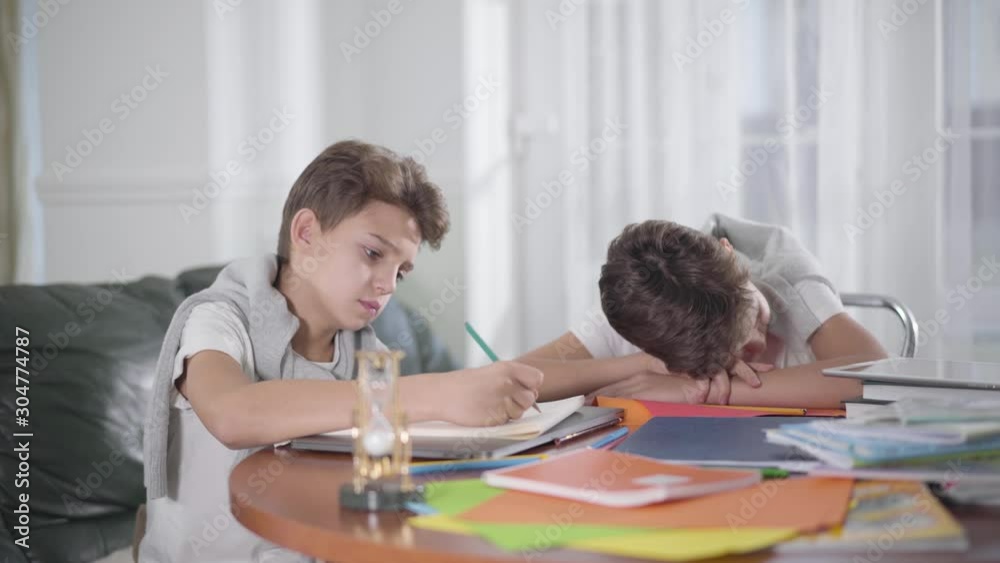 Side view of Caucasian schoolboy sitting at the table and writing down. His exhausted twin brother lying head on exercise book and falling asleep. Boy waking his sibling up and forcing him to study. Side view of Caucasian schoolboy sitting at the table and writing down. His exhausted twin brother lying head on exercise book and falling asleep. Boy waking his sibling up and forcing him to study.