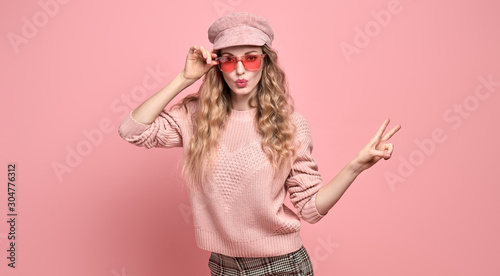 Easy-going Woman happy smiling in Stylish fashion pink colored outfit. Beautiful young Girl in Trendy jumper, make up. Joyful blonde lady shows peace sign in pink cap, fashionable happiness concept