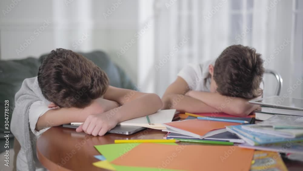 Young Caucasian brunette boy waking up and stretching. His brother or friend sleeping on the table. Siblings felled asleep on books after doing homework. Exhaustion, education concept. Young Caucasian brunette boy waking up and stretching. His brother or friend sleeping on the table. Siblings felled asleep on books after doing homework. Exhaustion, education concept.