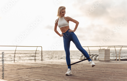 Healthy woman training on seaside promenade. Sportswoman in sportwear doing exercise with fitness rubber on the beach at sunrise