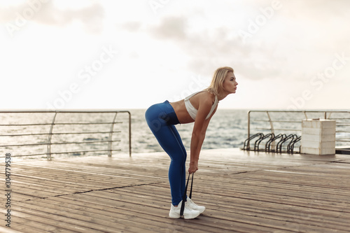 Healthy woman training on seaside promenade. Sportswoman in sportwear doing exercise with fitness rubber on the beach at sunrise