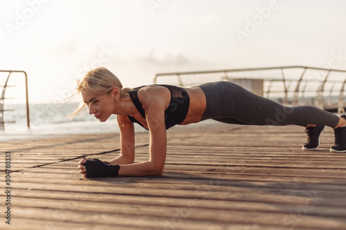 Healthy woman training on seaside promenade. Young sport woman in sportswear doing plank exercise on the beach at sunrise