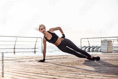 Healthy woman training on seaside promenade. Young sport woman in sportswear doing side plank exercise on the beach at sunrise