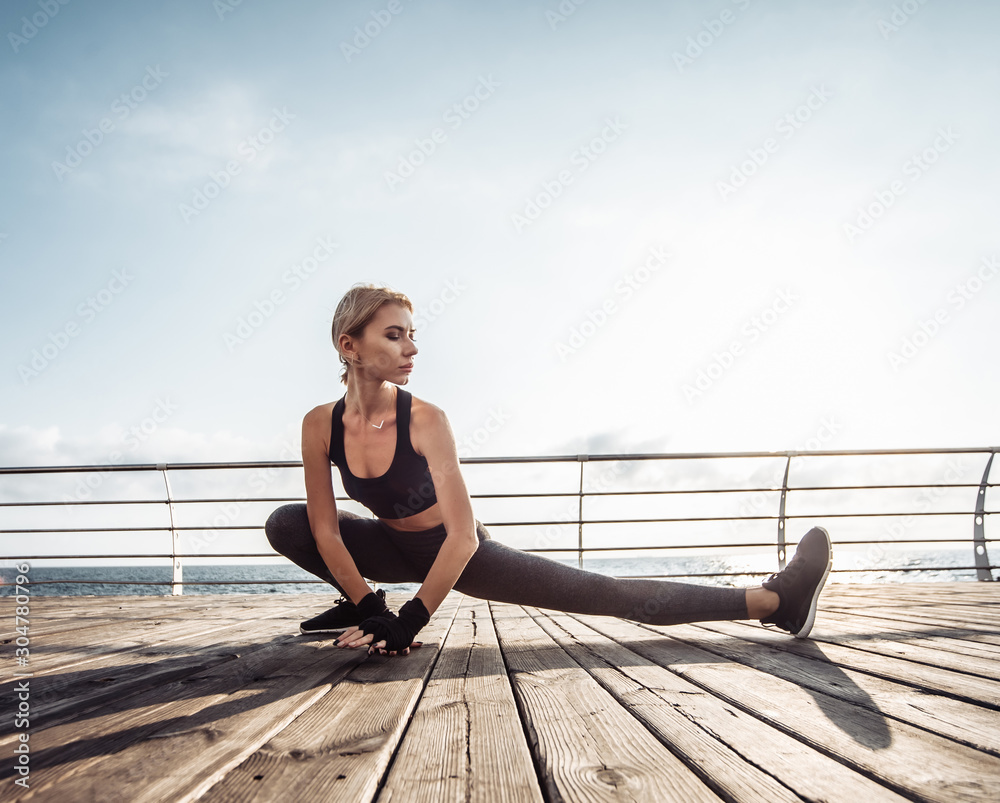 Healthy woman training on seaside promenade. Young attractive woman in sportswear does leg stretching before exercise on the beach at sunrise. Warm up