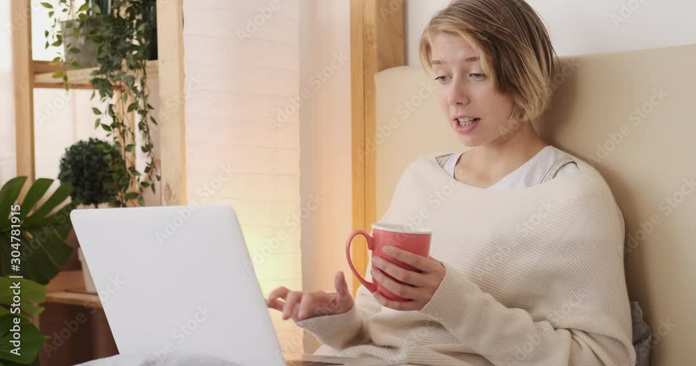 Young woman holding coffee mug while video chatting using laptop on bed Young woman holding coffee mug while video chatting using laptop on bed