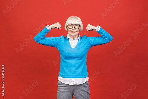 Healthy lifestyle. Happy elderly senior aged woman showing her muscle strong arms. Isolated on red background.