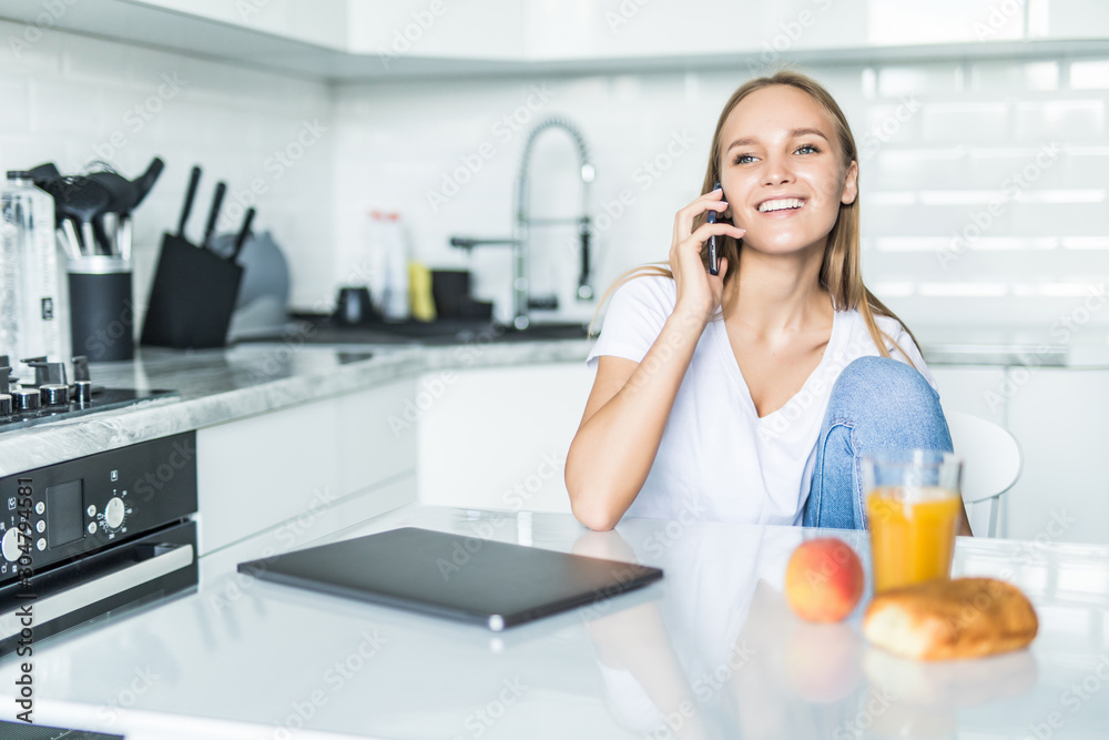 Young woman talking on mobile phone while using laptop in kitchen at home Young woman talking on mobile phone while using laptop in kitchen at home