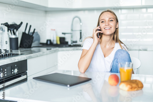 Young woman talking on mobile phone while using laptop in kitchen at home