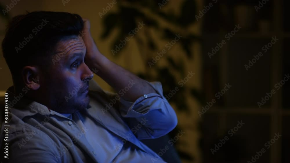 Chest up shot of upset Caucasian man crying and wiping tears with hand while watching dramatic movie on TV in dark living room in the evening