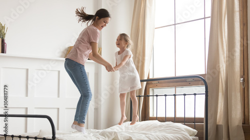 Playful mom have fun jumping on bed with little daughter