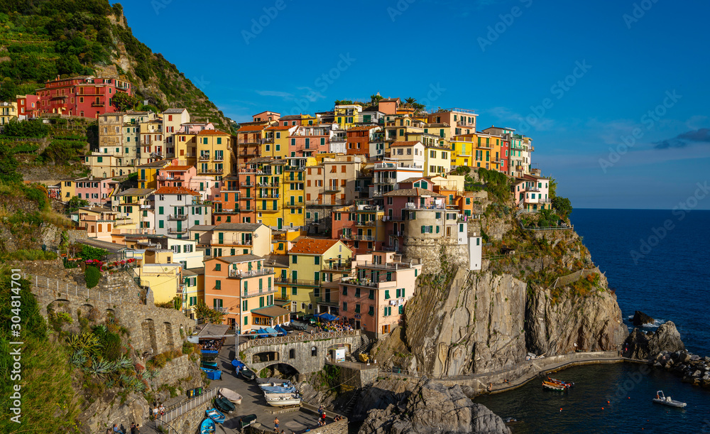 Manarola typical Italian village in National park Cinque Terre, colorful multi colored buildings houses on rock cliff, fishing boats on water, blue sky background, Liguria, Italy