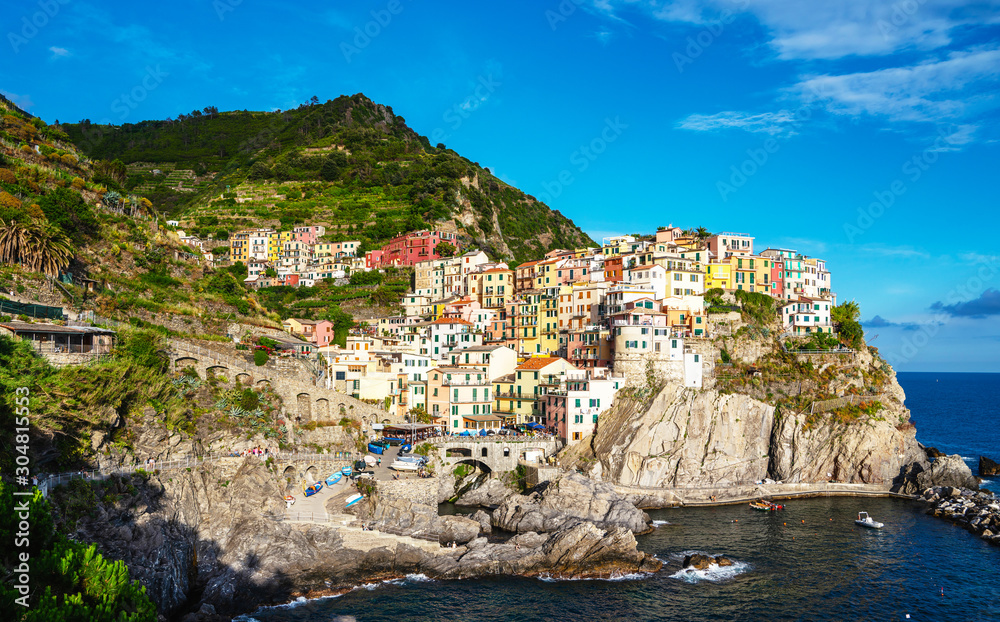 Manarola typical Italian village in National park Cinque Terre, colorful multi colored buildings houses on rock cliff, fishing boats on water, blue sky background, Liguria, Italy