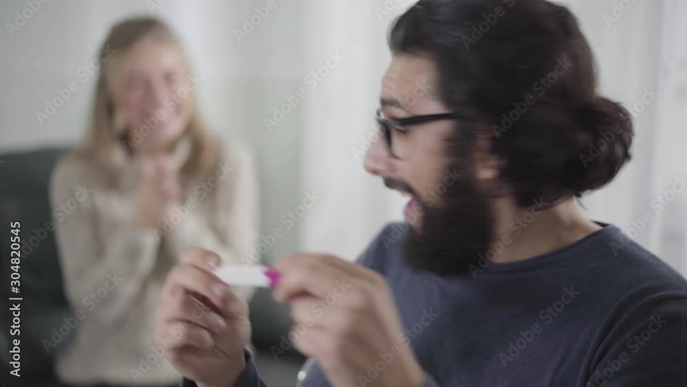 Close-up face of Caucasian man holding pregnancy test, looking back at his blond wife clapping hands, and smiling. Bearded guy in eyeglasses happy to become future father. Close-up face of Caucasian man holding pregnancy test, looking back at his blond wife clapping hands, and smiling. Bearded guy in eyeglasses happy to become future father.