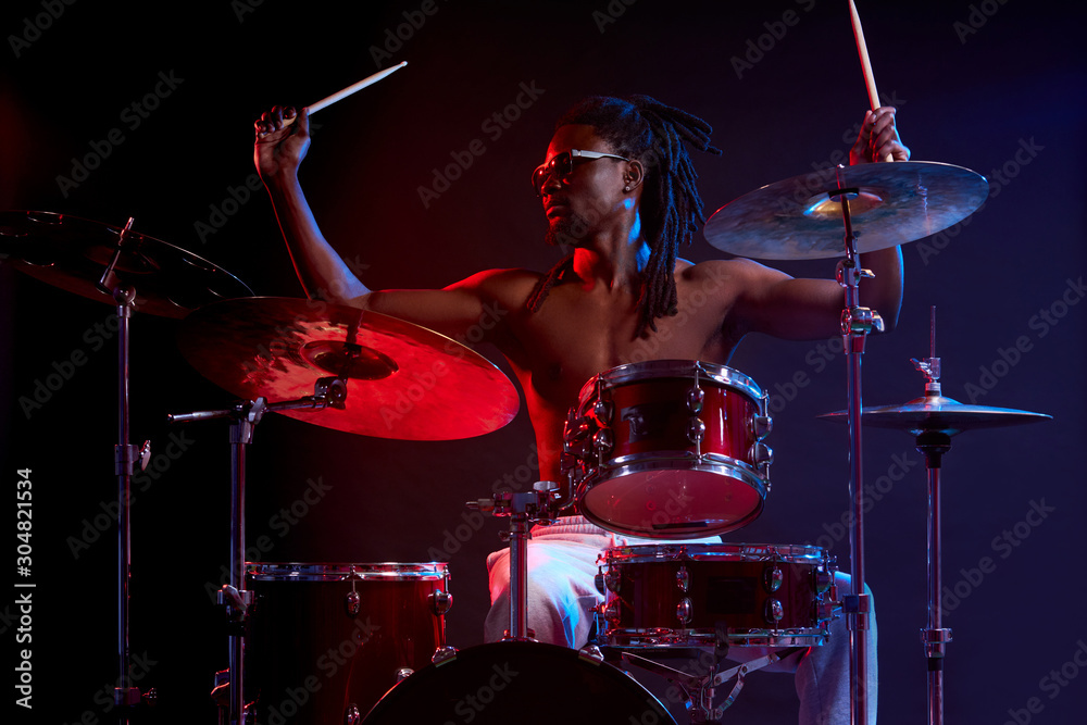 Fototapeta premium Portrait of active african man playing on drums set in neon lights, wearing eyeglasses, performing music. expressive emotions and movement