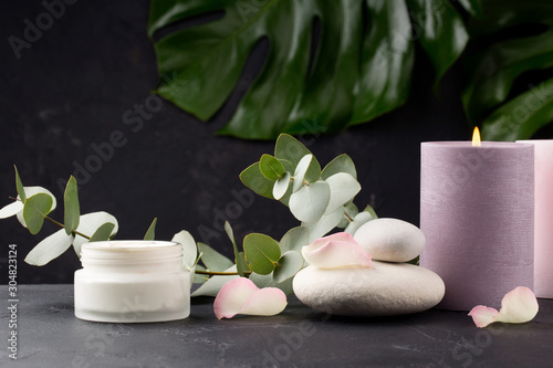 Spa concept. Cream with coconut milk, sea stones, eucalyptus and rose petals on black background. Copy space.