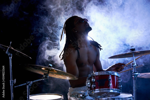 african young naked handsome man hitting on drums, perussion instruments isolated over smoky background. sit with head up, with dreadlocks