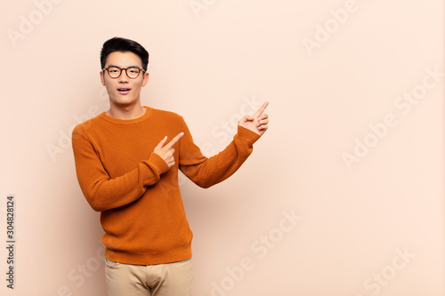 young chinese man feeling joyful and surprised, smiling with a shocked expression and pointing to the side against flat color wall