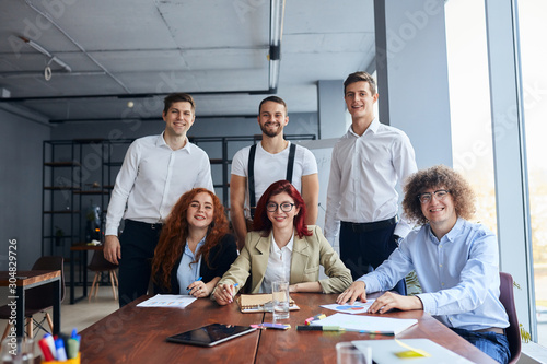 Smiling business team co-working sitting on table together and look at camera, wearing formal wear.