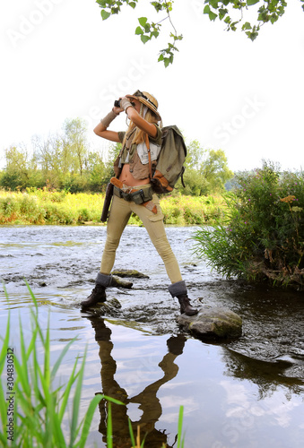 Fototapeta Naklejka Na Ścianę i Meble -  A young girl dresses up as an explorer. She wears a safari hat and wears khaki clothing. She is seen in a  river environment.