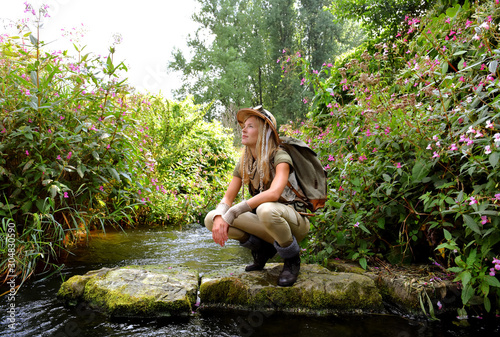A young girl dresses up as an explorer. She wears a safari hat and wears khaki clothing. She is seen in a  river environment.