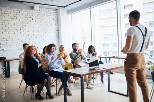 Confident enthusiastic young coach give speech to co-workers attentively listening to him in office, wearing casual wear