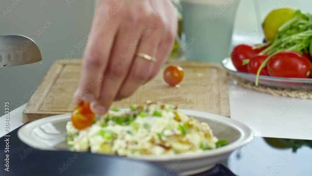 Close up of hands of unrecognizable male cook slicing cherry tomatoes and adding them to scrambled eggs with spring onion while preparing breakfast in kitchen Close up of hands of unrecognizable male cook slicing cherry tomatoes and adding them to scrambled eggs with spring onion while preparing breakfast in kitchen