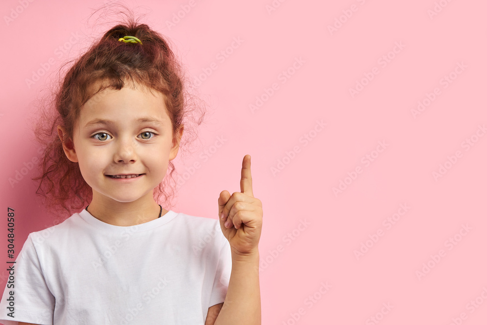 Smiling attractive girl kid show forefinger, wearing white t-shirt, isolated pink background Smiling attractive girl kid show forefinger, wearing white t-shirt, isolated pink background