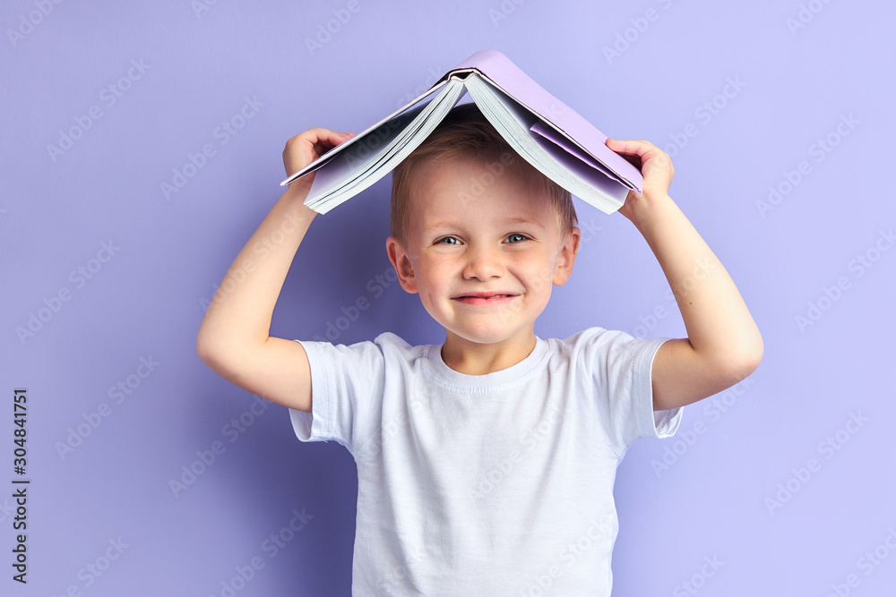 Portrait of joyful caucasian child in casual clothes after education, after reading wants to play. Funny boy holding book above his head, looking at camera Portrait of joyful caucasian child in casual clothes after education, after reading wants to play. Funny boy holding book above his head, looking at camera