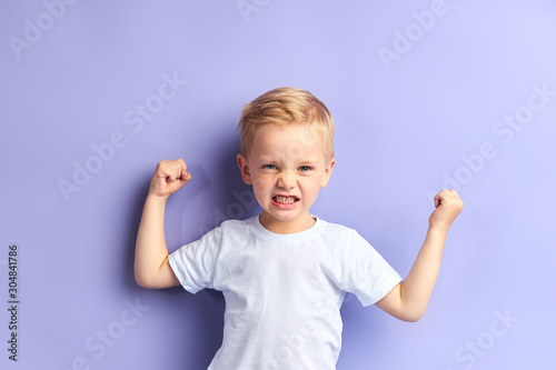Portrait of caucasian kid boy with blond hair showing muscles at camera, wearing white t-shirt. Purple background