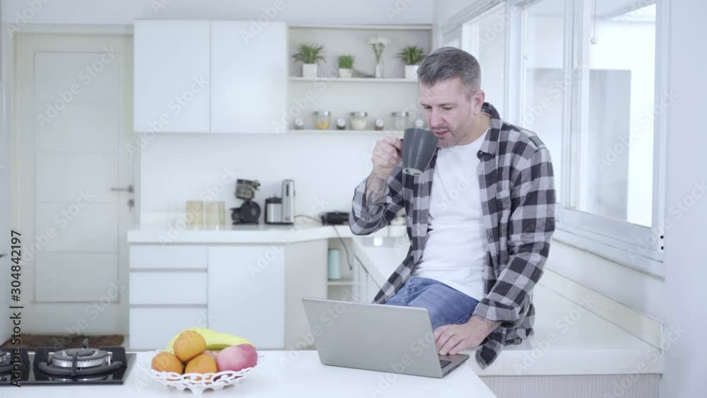 Handsome European man using a laptop computer while drinking a cup of coffee in the kitchen at home. Shot in 4k resolution Handsome European man using a laptop computer while drinking a cup of coffee in the kitchen at home. Shot in 4k resolution