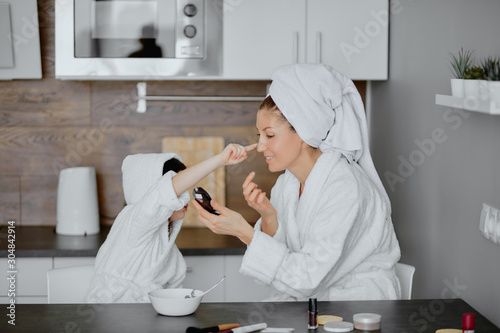 cute child touches mother's nose, using facial mask. sit toether in kitchen wearing bathrobes and towels photo