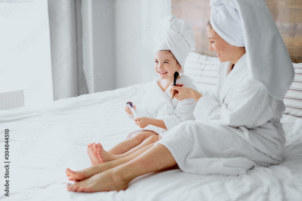 Fototapeta premium caucasian woman and little daughter sit on bed wearing towels and bathrobe doing makeup after shower. family concept